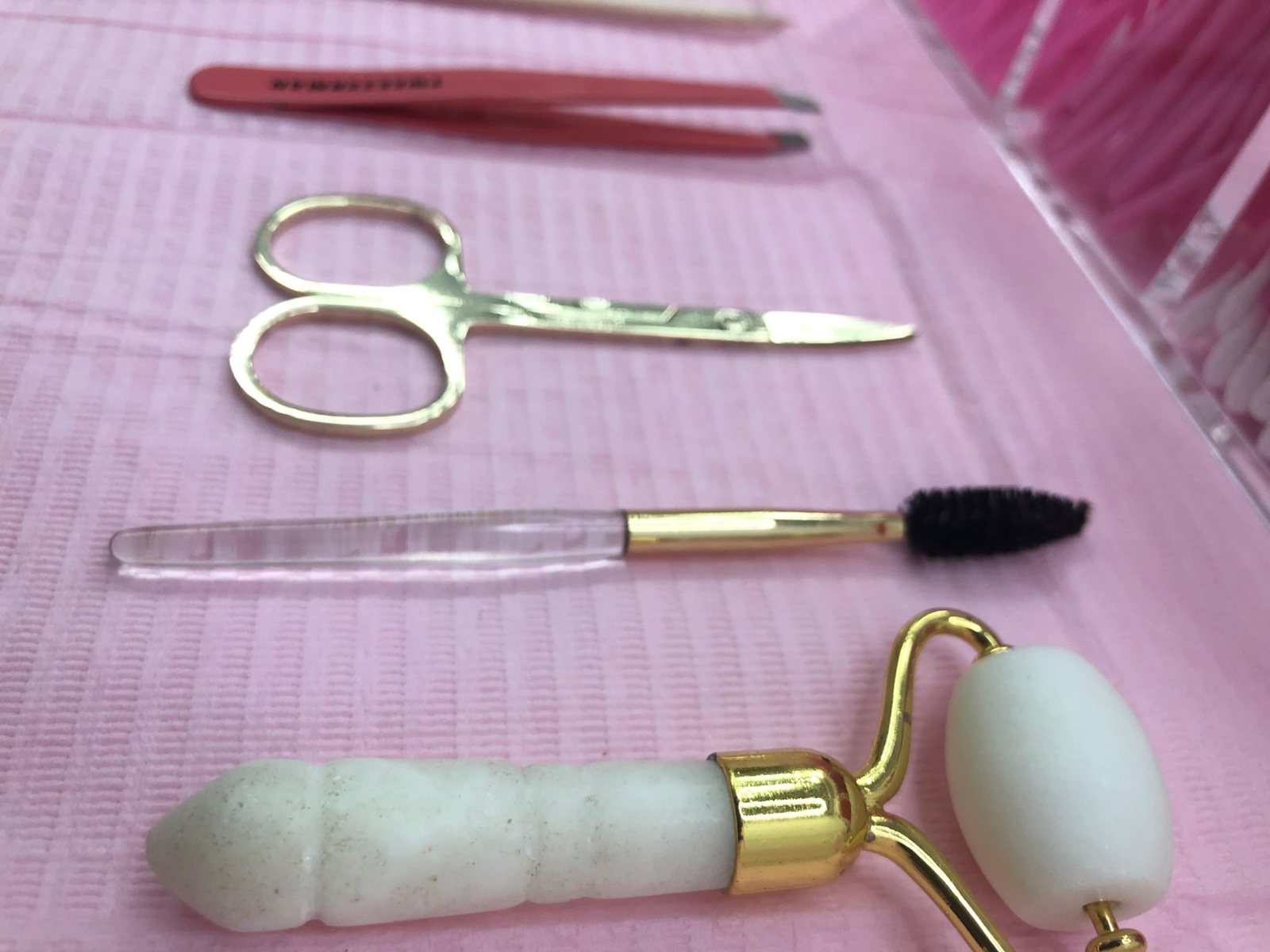 Tweezers, scissors, a spoolie and a face roller laid out on pink
